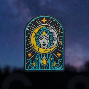 May include: Embroidered patch featuring a celestial design. The patch showcases a woman's face within a crescent moon, with teal hair and accents. The design includes stars and a teardrop, all set against a black background with a teal border.
