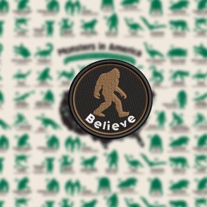 May include: A round patch featuring a brown Bigfoot silhouette walking to the left, with the word "Believe" embroidered in white. The patch has a black border with a brown inner ring. The background is a cream color with green monster illustrations.