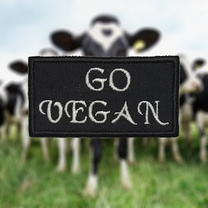 May include: Black embroidered patch with white text that reads "GO VEGAN" against a blurred background of cows.