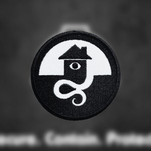 May include: Black and white embroidered patch with a house design and an eye in the window. The house is surrounded by a black and white swirl.