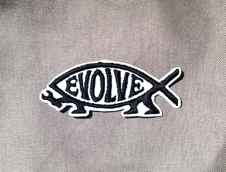 May include: Black and white embroidered patch of a fish with the word "EVOLVE" inside. The patch is on a gray fabric background.