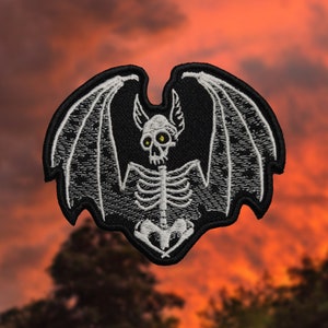 Puede incluir: Parche de esqueleto de murciélago bordado en blanco con alas negras y ojos amarillos. El parche está sobre un fondo de puesta de sol rojo y naranja.