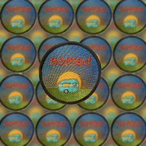 May include: A close-up of a colourful embroidered patch with the word "Nomad" in red lettering. The patch features a sunset scene with a blue camper van in the foreground.