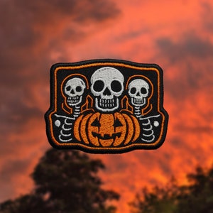 May include: Embroidered patch featuring three white skulls and an orange jack-o'-lantern on a black background. The patch has an orange border and is designed for Halloween. The skulls have detailed features, and the pumpkin has a carved face.