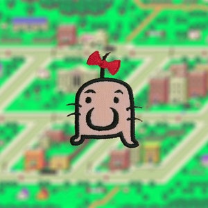 May include: Embroidered patch of a pink cartoon character with a red bow on its head. The character has a black outline and a smiling face.