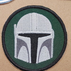 May include: Embroidered patch featuring a Mandalorian helmet design. The helmet is primarily white and gray, with a black visor and accents. The patch is circular, with a green background and a black border. The design is detailed and appears to be machine-embroidered.