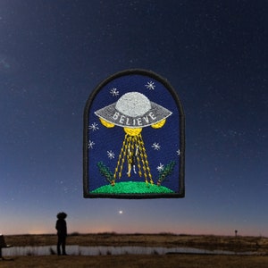 Peut inclure: Un patch brodé bleu avec un OVNI blanc sur lequel est inscrit le mot "BELIEVE". L'OVNI est entouré d'étoiles et des rayons de lumière jaunes en émanent.