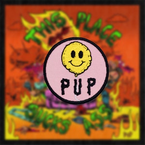 Puede incluir: Un parche bordado rosa y amarillo con un globo sonriente y la palabra "PUP" en letras negras. El parche está sobre un fondo de una imagen de dibujos animados con las palabras "This Place Sucks" en letras verdes y naranjas.
