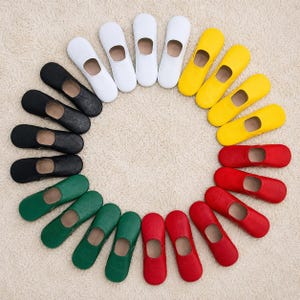 May include: A circular arrangement of nineteen slip-on slippers in various colors, including white, yellow, red, green, and black. Each slipper features a rounded toe, a low-cut opening, and a smooth, solid-colored exterior finish.