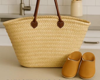 Op de afbeelding: Een geweven strooien draagtas met bruine leren handvatten en accenten, gecombineerd met een paar bruinleren Marokkaanse slippers. De tas heeft een breed, open ontwerp. De slippers zijn effen.
