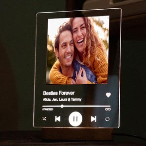 以下が含まれることがあります： 2人の笑顔の写真が入った透明アクリルフォトフレーム。「Besties Forever」の文字と名前が書かれた音楽プレーヤーのインターフェースデザイン。日付07/04/2021が表示されています。