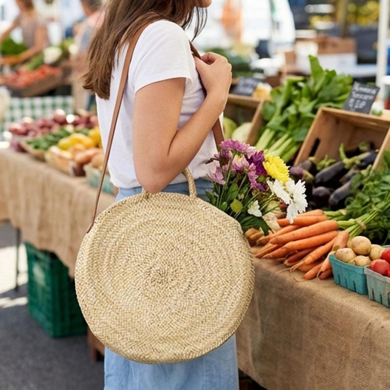 Op de afbeelding: Een ronde, geweven strooien schoudertas met een bruine leren riem. De tas wordt door een persoon op een markt gedragen, gevuld met bloemen. De marktkraam toont verse producten, waaronder wortelen en aubergines.