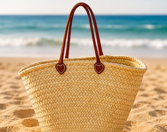 May include: A woven straw basket tote bag with a wide, open top and two long, brown leather handles. The handles are attached to the body of the bag with heart-shaped leather patches featuring white stitching at the base of each strap.