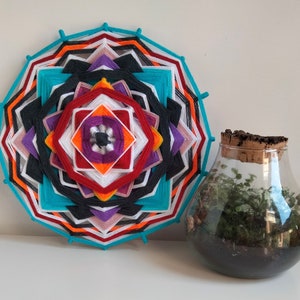 Peut inclure: Une tapisserie murale mandala colorée avec un motif géométrique dans des tons de rouge, orange, jaune, blanc, noir, violet et bleu. La mandala est faite de fil et est accrochée à un mur blanc. Un terrarium en verre avec un couvercle en liège et une petite plante à l'intérieur est posé sur une surface blanche devant la mandala.