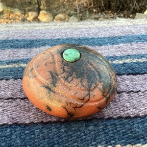 May include: A round, orange-colored ceramic dish with black and gold swirling patterns. The dish has a small, round, turquoise-colored stone in the center.