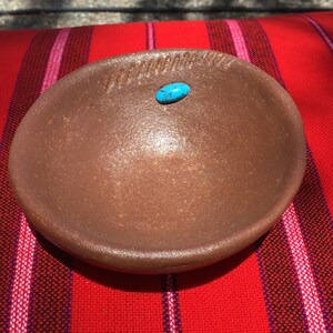 May include: A small, brown, ceramic bowl with a turquoise stone in the center. The bowl is sitting on a red and white striped blanket.