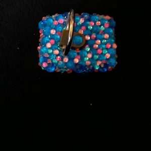 May include: A small, square-shaped, blue and pink jeweled keychain with a silver ring.