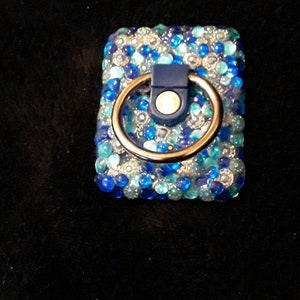 May include: A rectangular phone grip with a ring holder, covered in blue and clear rhinestones. The ring holder is gold-colored, and the base is navy blue. The phone accessory is designed to attach to the back of a phone.