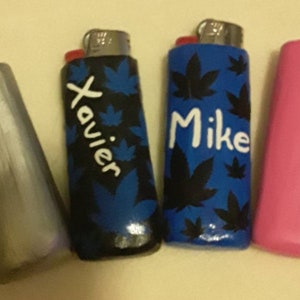 May include: Four lighters with personalised names. Two lighters have a blue background with black leaf designs and white lettering. One lighter has a black background with blue leaf designs and white lettering. One lighter is pink.