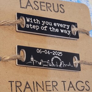 May include: Two black metal dog tags with white text and a small string attached. The top tag reads "With you every step of the way". The bottom tag has a silhouette of the London skyline with the date "06-04-2025" and the text "TRAINER TAGS".