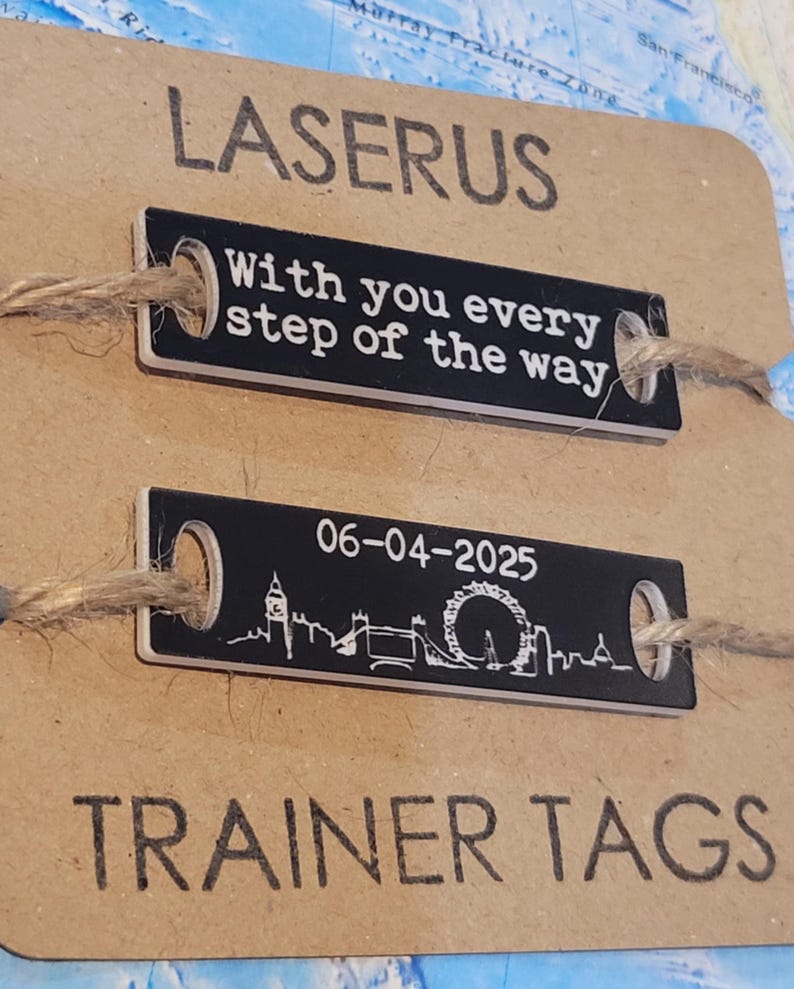 May include: Two black metal dog tags with white text and a small string attached. The top tag reads "With you every step of the way". The bottom tag has a silhouette of the London skyline with the date "06-04-2025" and the text "TRAINER TAGS".
