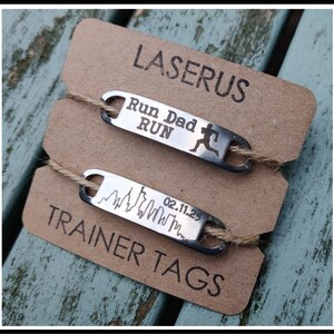 May include: Two silver metal tags with engraved text. The top tag reads "Run Dad Run" with a running figure. The bottom tag reads "02.11.25" with a skyline graphic. Both tags are on a brown card with the text "Laserus Trainer Tags".