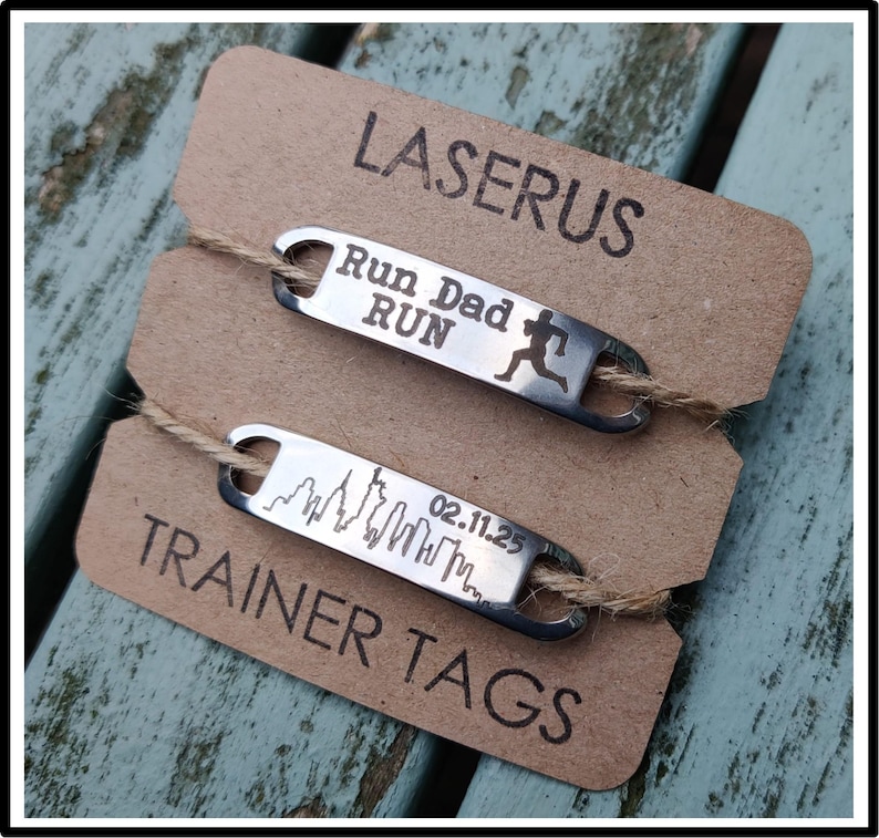 May include: Two silver metal tags with engraved text. The top tag reads "Run Dad Run" with a running figure. The bottom tag reads "02.11.25" with a skyline graphic. Both tags are on a brown card with the text "Laserus Trainer Tags".