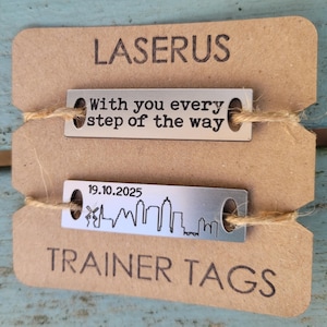 May include: Two silver metal tags with a brown paper background. The top tag reads "With you every step of the way". The bottom tag reads "19.10.2025" and has a skyline silhouette. The text "LASERUS TRAINER TAGS" is printed on the paper background.