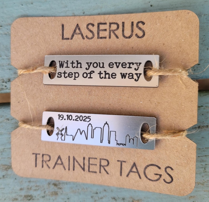 May include: Two silver metal tags with a brown paper background. The top tag reads "With you every step of the way". The bottom tag reads "19.10.2025" and has a skyline silhouette. The text "LASERUS TRAINER TAGS" is printed on the paper background.