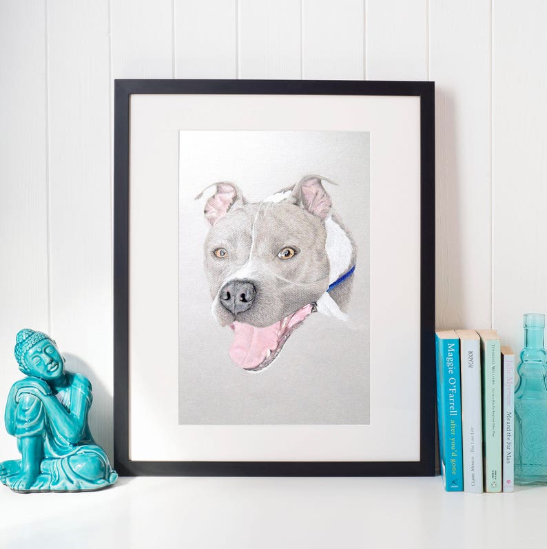 Op de afbeelding: Een ingelijst portret van een hond. De afbeelding toont een close-up van de kop en schouders van de hond, met grijs en wit haar, een roze tong en een blauwe halsband. Het kunstwerk zit in een zwarte lijst met een witte passe-partout.