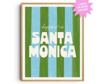 May include: A framed art print with a light wood frame. The artwork features vertical stripes in alternating shades of blue and green. The words "happiest in SANTA MONICA" are in white, bold font. A pink starburst graphic says "COMES IN MORE COLORS".