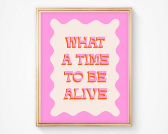 May include: A framed art print with the words "WHAT A TIME TO BE ALIVE" in a retro font. The text is in orange and pink, set against a cream background with a wavy pink border, all within a gold frame. The artwork is a vibrant and positive statement piece.