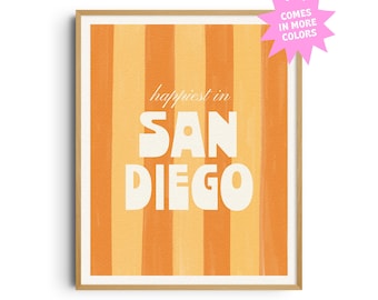 May include: A framed art print with a vertical striped background in shades of orange and yellow. The text "happiest in SAN DIEGO" is displayed in a bold, white font. A pink starburst graphic indicates the availability of more colors.