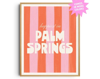 May include: A framed art print with a vertical striped pattern in shades of pink and orange. The words "happiest in PALM SPRINGS" are displayed in white, bold lettering. A pink starburst graphic states "COMES IN MORE COLORS".