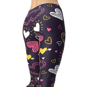 May include: Black yoga leggings with a colourful heart pattern. The hearts are in shades of pink, yellow, and white. The leggings are full length and have a tight fit.