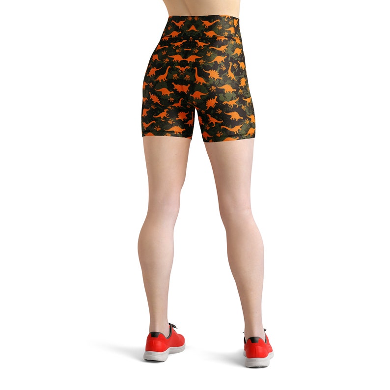 May include: Black and green high-waisted shorts with a repeating pattern of orange dinosaurs. The shorts are worn by a person with bare legs and red sneakers.