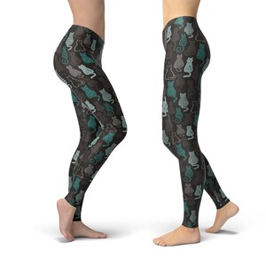 May include: Black leggings with a repeating pattern of stylized cats in teal and gray. The cats have various designs, including stripes and solid fills. The leggings are shown from the knees down, with a white background.