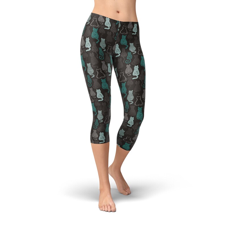 May include: Cropped leggings with a repeating pattern of stylized cats in teal, gray, and white on a dark gray background. The leggings are mid-calf length and form-fitting, suitable for athletic or casual wear.