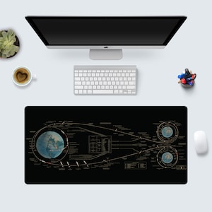 Op de afbeelding: Een zwarte bureaumat met een gedetailleerd diagram van de aarde en de ruimte, met een gouden ontwerp. Het diagram bevat lijnen, cirkels en tekst.