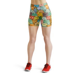 May include: A pair of athletic shorts with a colorful print of cartoon cats. The shorts are a bright yellow, green, blue, and orange. The model is wearing red athletic shoes.
