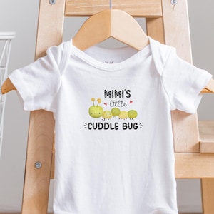 Puede incluir: Un body blanco para bebé con mangas cortas y un gráfico de una oruga verde con el texto "Mimi's little cuddle bug".