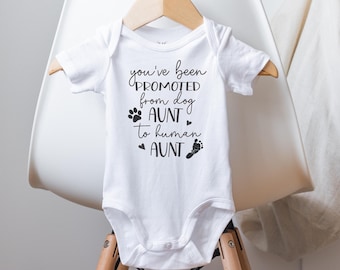 Może przedstawiać: Biały śpioszek dla niemowlęcia z czarnym napisem "You've been promoted from dog aunt to human aunt". Śpioszek wisi na drewnianym wieszaku na białym krześle.