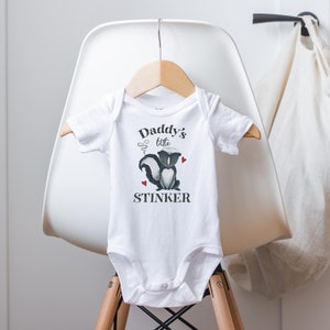 May include: White baby onesie with a black and white skunk illustration and the text "Daddy's little stinker".