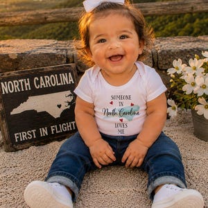 May include: A white short-sleeved bodysuit with the text "SOMEONE IN North Carolina LOVES ME" and a small map graphic, paired with dark blue jeans, white sneakers, and a white hair bow. A black sign with "NORTH CAROLINA FIRST IN FLIGHT" is nearby.
