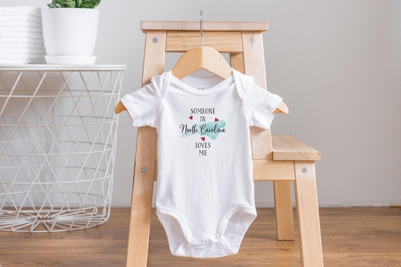 May include: A white short-sleeved baby bodysuit featuring the black text 'SOMEONE IN North Carolina LOVES ME' with a light blue silhouette of the state of North Carolina and three small red hearts printed on the front center of the garment.