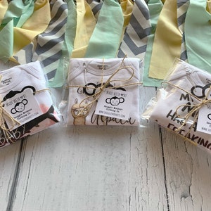 May include: Three white baby bodysuits packaged in clear plastic, each tied with brown twine and a square tag reading Made with by Snuggle Monkeys Clothing Co. The garments feature black and brown graphic prints of animals and text on the front.