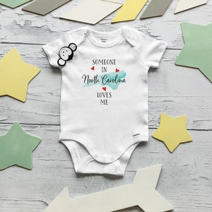 May include: A white short-sleeved baby bodysuit featuring the black text "SOMEONE IN North Carolina LOVES ME" with a light blue watercolor map of North Carolina and three small red hearts printed on the front chest area.