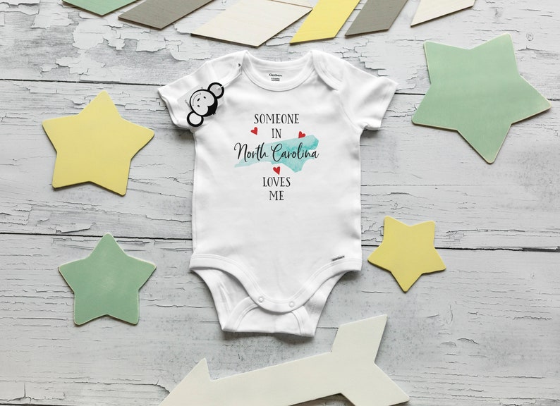 May include: A white short-sleeved baby bodysuit featuring the black text "SOMEONE IN North Carolina LOVES ME" with a light blue watercolor map of North Carolina and three small red hearts printed on the front chest area.