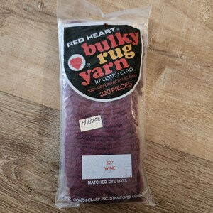 May include: A package of Red Heart Bulky Rug Yarn in the color wine, made of 100% Orlon acrylic fiber. The clear plastic packaging features a black label with red and yellow text, including the label 320 PIECES and a handwritten HB100 tag.