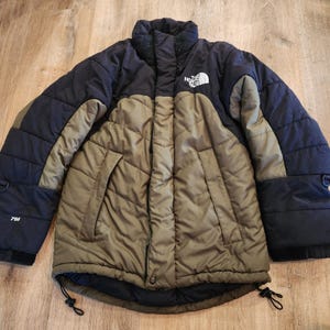 May include: A black and olive green puffer jacket featuring a high collar, a front zipper closure, and two side pockets. The North Face logo is embroidered in white on the upper chest, and the number 700 is printed on the right sleeve.